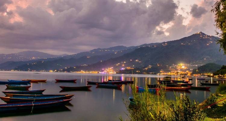 Coloridas barcas de madera flotan en un lago tranquilo al atardecer con luces titilantes en las laderas y siluetas de montañas de fondo.