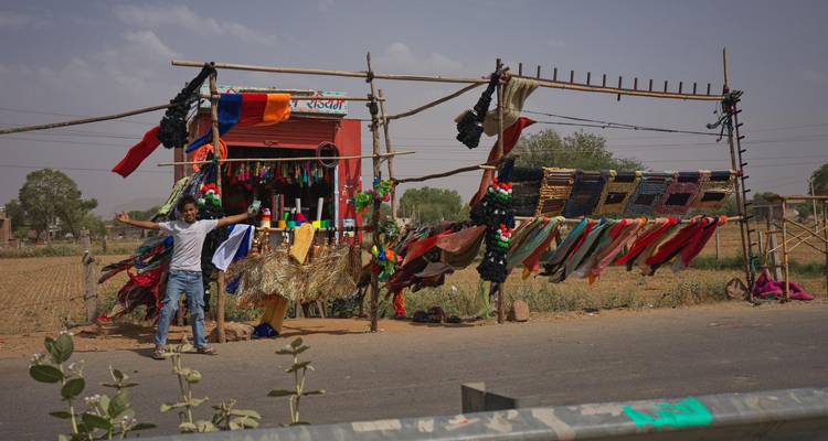 Petit étalage de tissus en bordure de route dans le Rajasthan rural avec des foulards colorés suspendus à des perches en bambou et un vendeur souriant.