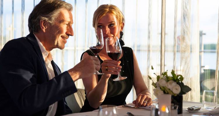 Ein elegantes Paar stößt während eines Candlelight-Dinners neben einem Fenster mit Wasserblick mit Weingläsern an.