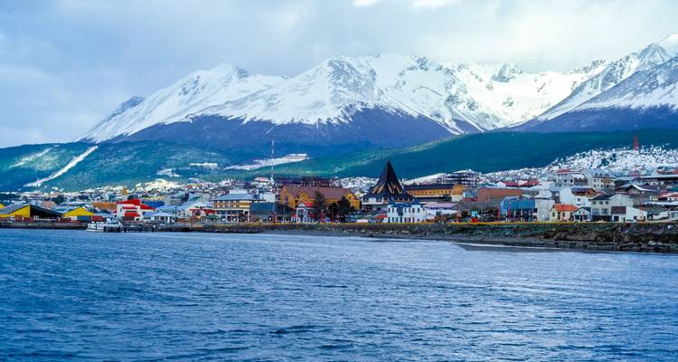 Küstenstadt Ushuaia mit schneebedeckten Andenspitzen im Hintergrund und tiefblauem Kanal im Vordergrund