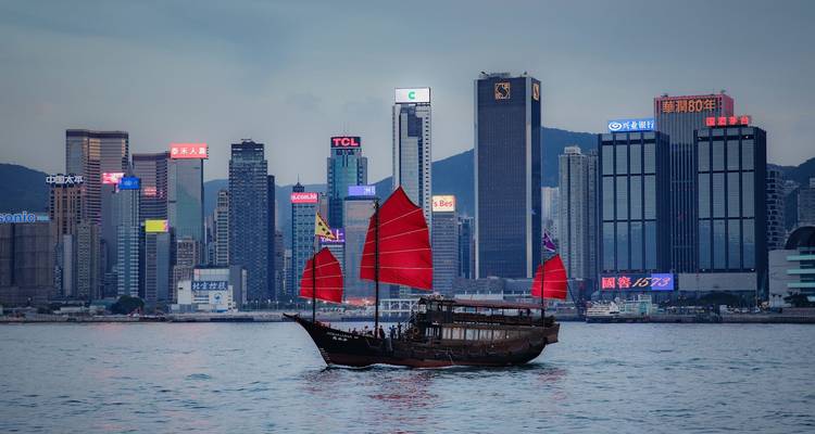 Un barco junk tradicional con velas rojo brillante se desliza por el Puerto Victoria con los rascacielos modernos de Hong Kong de fondo.