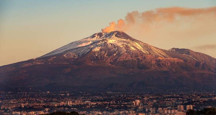 Rokende top van de met sneeuw bestoven Mount Etna domineert tijdens het gouden uur de skyline boven de stad.