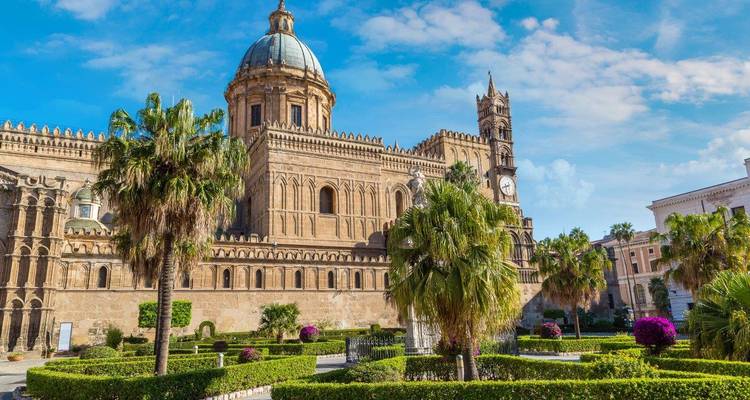Voorkant van de Kathedraal van Palermo omgeven door verzorgde tuinen en palmbomen onder een blauwe lucht.