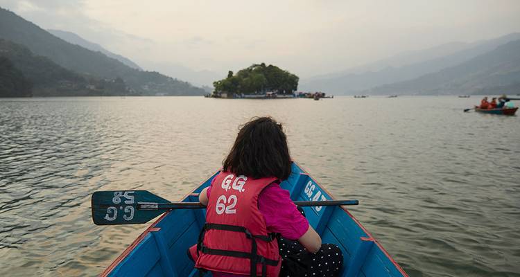 Woman paddles a blue wooden boat on a calm lake surrounded by misty hills at dusk.