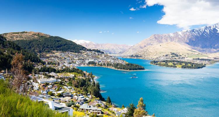Panorama costero brillante de los suburbios de Queenstown abrazando la curva del lago turquesa bajo picos áridos.