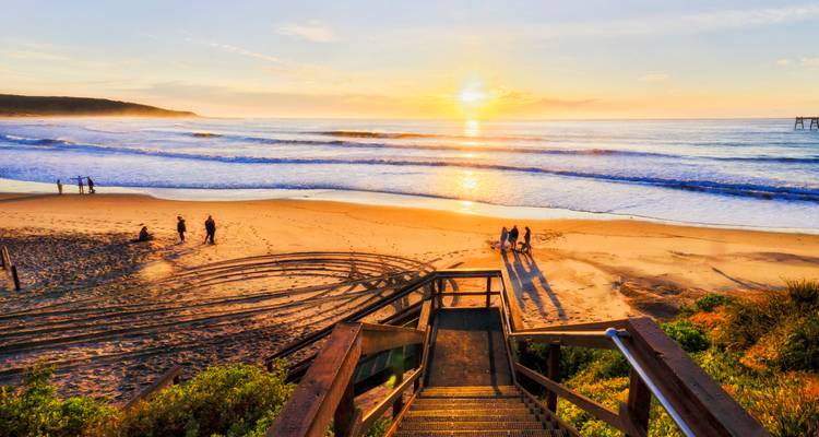 Lever de soleil doré sur une plage de sable avec de petits groupes se promenant près des vagues douces et des escaliers d'accès en bois au premier plan.