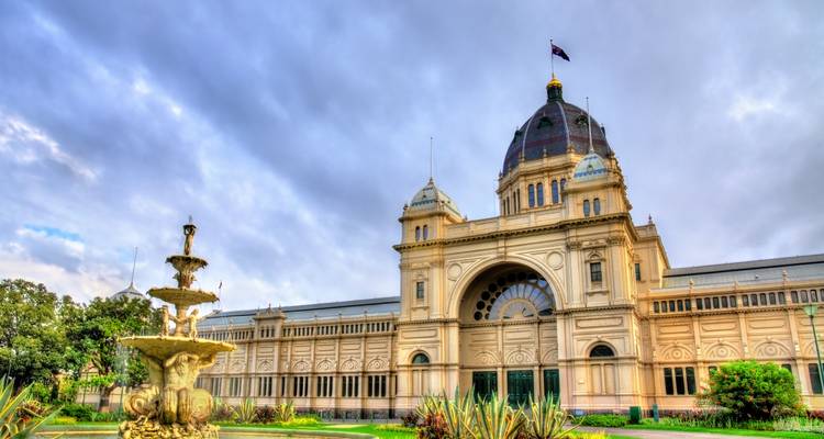 Bâtiment de l'Exposition Royale à Melbourne avec fontaine ornée et jardins paysagers sous un ciel nuageux.