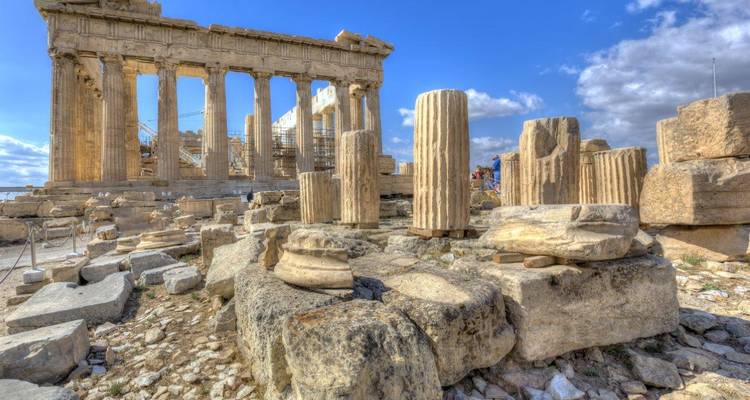 Close-up van Parthenon-zuilen en verspreid liggende marmeren blokken onder een strakblauwe lucht.
