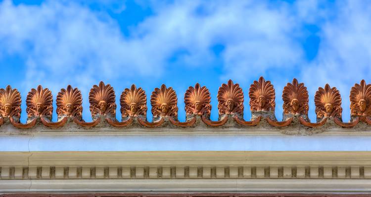 Decoratieve terracotta antefixen langs de rand van een klassiek dak tegen een helderblauwe hemel.