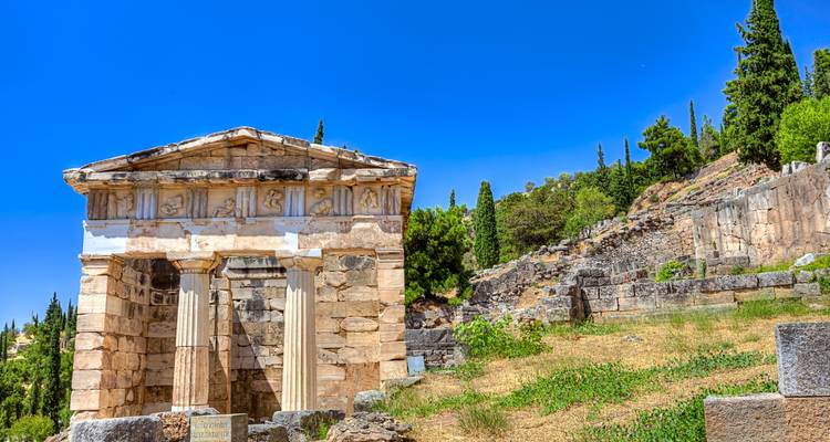 Gereconstrueerd Athenaans Schatkuis in Delphi met levendige blauwe lucht en ruïnes eromheen.