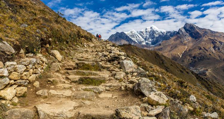 El sendero de escaleras de piedra sube por una cresta escarpada hacia una cumbre nevada bajo un cielo salpicado de nubes.