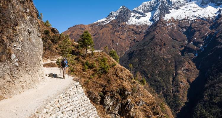 Un trekker en solitario camina por un estrecho sendero de montaña cortado en acantilados escarpados con un valle boscoso abajo.