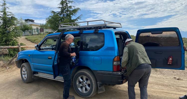 Two men load equipment into a blue 4x4 parked on a dirt road under patchy clouds.