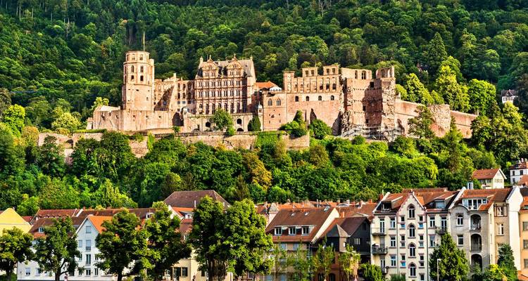 Primer plano de las torres y muros del Castillo de Heidelberg emergiendo de un denso follaje verde por encima de las casas del pueblo.