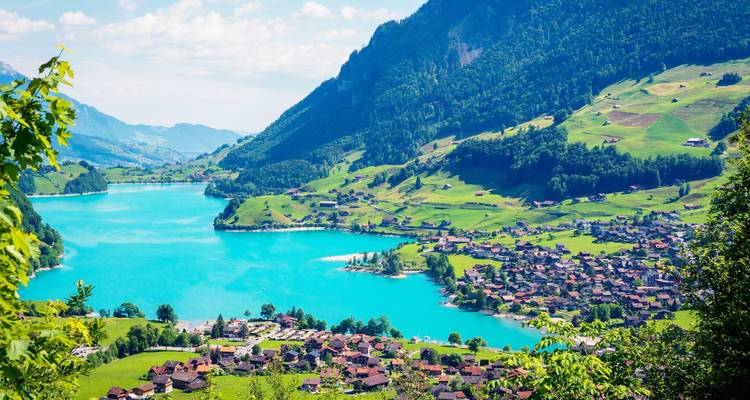 Un lago alpino de un turquesa vibrante rodeado de colinas verdes ondulantes y pueblos suizos tradicionales bajo un cielo brillante.