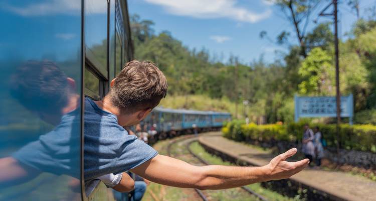 Viajero asomándose de un tren azul de Sri Lanka con el brazo estirado mientras los vagones se deslizan por las curvas de las vías de la selva.