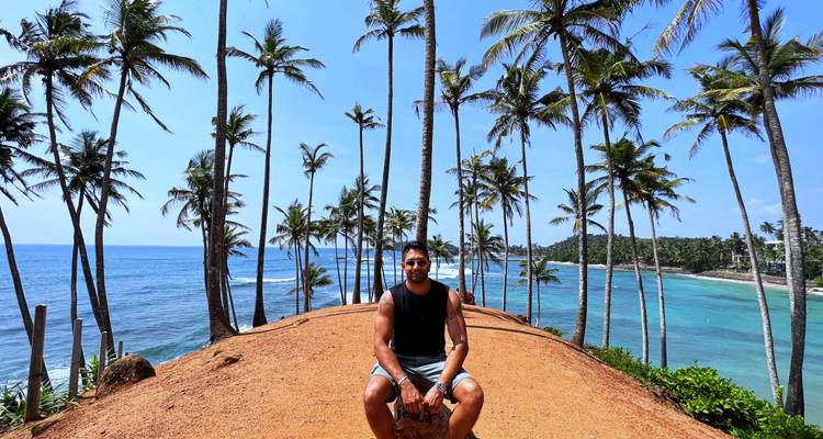 Un hombre sentado en una cresta de tierra roja rodeada de altos cocoteros con vistas al mar turquesa en Coconut Tree Hill.