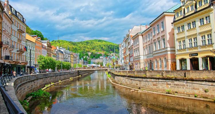 Canal pintoresco flanqueado por coloridos edificios históricos de balneario en Karlovy Vary con colinas verdes más allá.