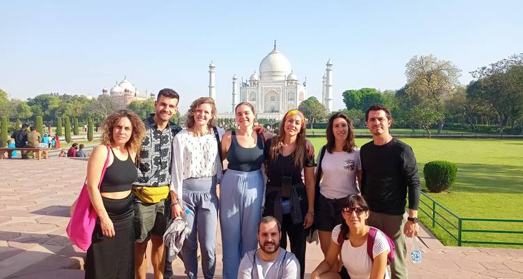Eine lächelnde Reisegruppe posiert zusammen mit dem glänzenden Taj Mahal im Hintergrund an einem sonnigen Morgen.