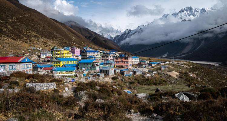 Vista panorámica de un pueblito del Himalaya pintado con colores brillantes, acurrucado contra las laderas empinadas con nubes envolviendo las cimas nevadas.