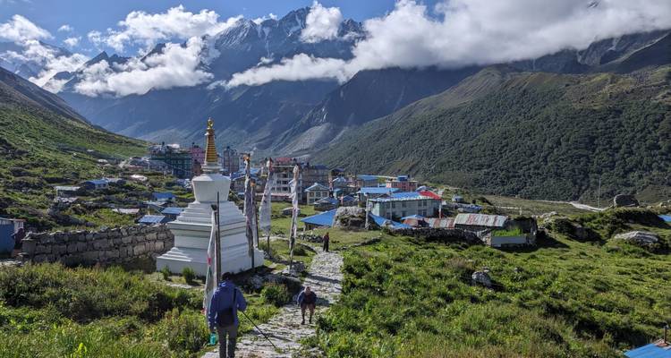 Un estupa blanco y un camino de piedra guían a los trekkers hacia un pueblo de montaña, enmarcado por colinas exuberantes y picos coronados de nubes.