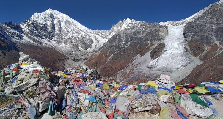 Coloridas banderas de oración ondean en un mirador rocoso que domina glaciares y escarpadas cimas del Himalaya bajo un cielo azul profundo.
