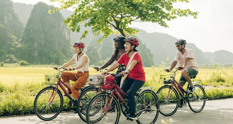 Quatre voyageurs circulent à vélo classique sur une route rurale ensoleillée encadrée par des pics karstiques.