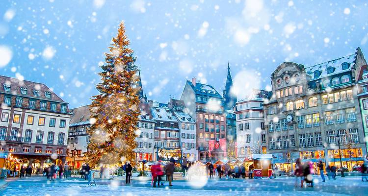 Snow falls over Strasbourg’s festive square with a giant Christmas tree and historic timbered buildings.