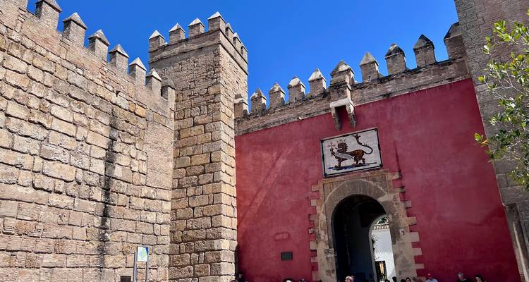 Mur rouge et porte en pierre crénelée de l'Alcázar de Séville avec des visiteurs à l'entrée.