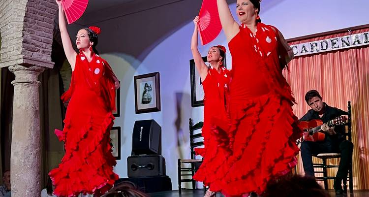 Des danseuses de flamenco vêtues de robes rouges éclatantes se produisent de manière dramatique sur scène, accompagnées par un guitariste.