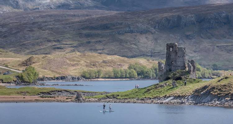 Dos practicantes de paddleboard se deslizan en un loch tranquilo junto al castillo en ruinas de Ardvreck bajo un cielo azul.