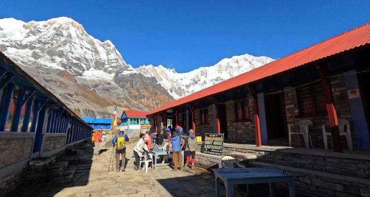 Trekkings verzamelen zich bij een stenen gasthuisvestiging met sneeuwbedekte Himalaya-toppen die in de achtergrond omhoog rijzen.