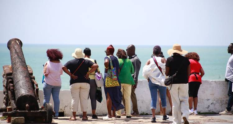 A tour group stands along fortress walls with an old cannon, looking out over the Atlantic Ocean.
