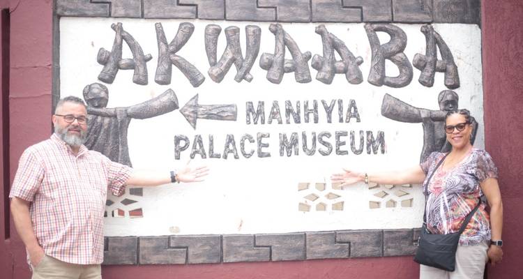 Two tourists point at the welcome sign for Manhyia Palace Museum with smiles on their faces.