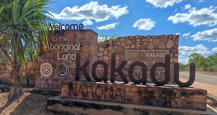 Letrero de bienvenida de piedra que dice "Kakadu National Park" bajo un cielo azul brillante punteado con nubes esponjosas.