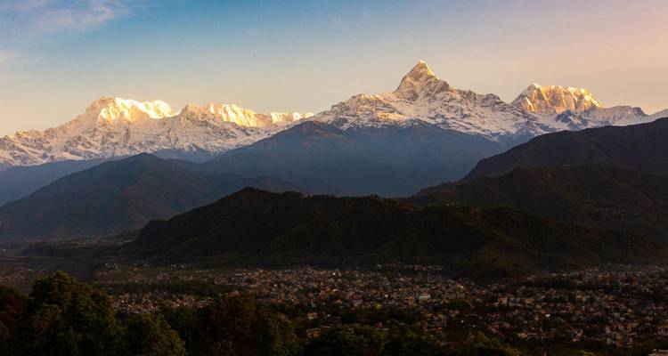 Des sommets de l'Himalaya enneigés brillant à la lumière du matin au-dessus d'une ville de vallée et de collines boisées.
