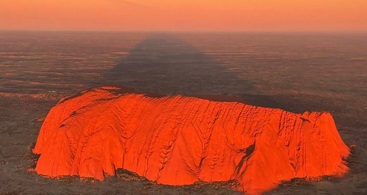 An aerial view of Uluru glowing deep orange in the setting sun, casting a vast shadow across the desert.