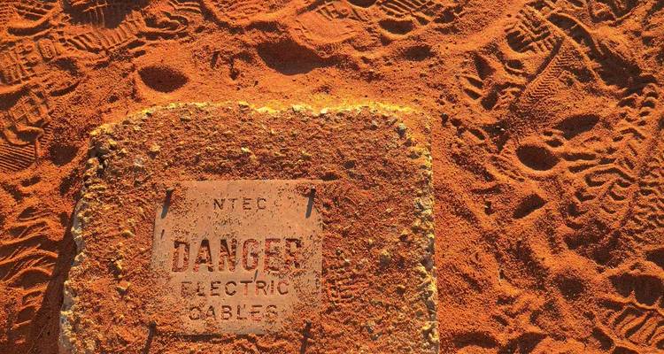 Close-up of a metal warning plate reading 'DANGER ELECTRIC CABLES' set in red desert sand with boot prints.