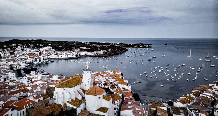 Aerial view of Cadaqués with white buildings and terracotta roofs hugging the coastline and marina.