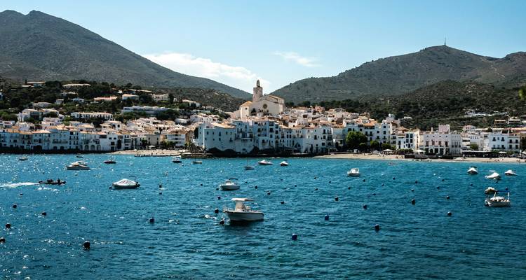 Blue waters of Cadaqués bay with the town and chapel framed by surrounding hills under a bright sky.