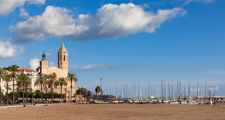 Historic church tower and marina masts rise above Sitges beachfront on a sunny day with blue skies.