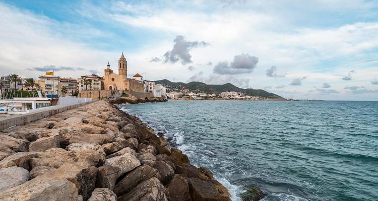 Rocky breakwater leading to the town of Sitges with church tower and coastal hills under partly cloudy skies.