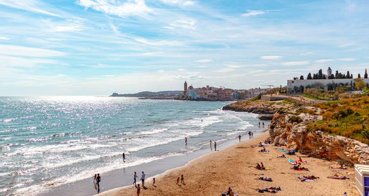 Sun-drenched beach at Sitges with people swimming and sunbathing, the old town visible in the distance.