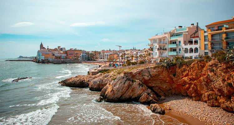 Coastal cliffs and colorful buildings of Sitges overlooking the Mediterranean with gentle surf below.