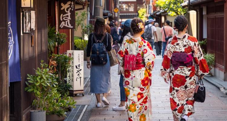 Zwei Frauen in farbenfrohen Kimonos spazieren durch eine traditionelle Holzgasse mit Geschaeften.