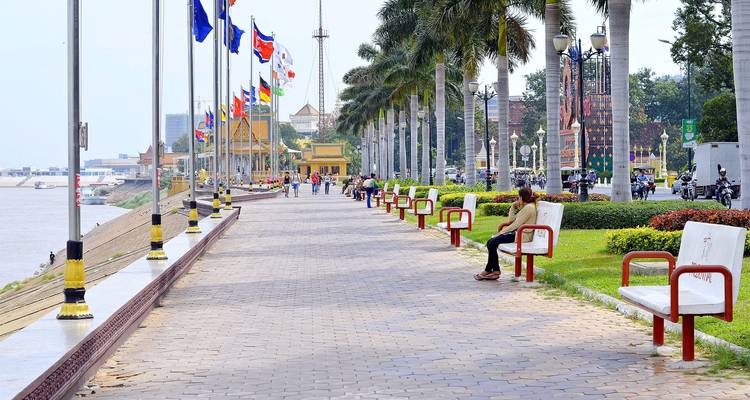 Lange rivierpromenade met palmbomen en kleurrijke internationale vlaggen, waar mensen op bankjes relaxen.