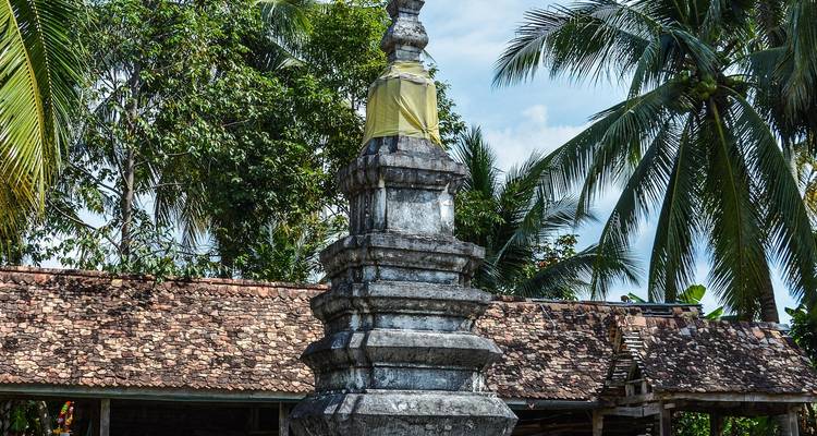 Kleine stenen stupa verpakt in geel doek omgeven door palmbomen en rustieke dakpannen.