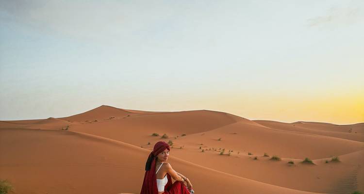 Una mujer sola con un pañuelo rojo en la cabeza se sienta en una enorme duna de arena naranja al atardecer en el Desierto del Sahara.
