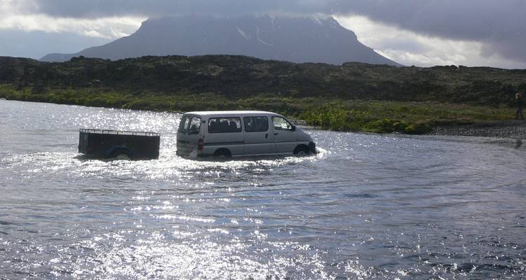 Van und Anhänger beim Durchfahren eines flachen Flusses vor der Kulisse eines Vulkanplateaus