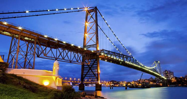 Puente colgante iluminado que atraviesa un canal tranquilo a la hora azul con las luces de la ciudad reflejándose en el agua